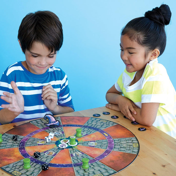 Two children play a colorful circular board game with dice and green potion-shaped pieces on a wooden table.