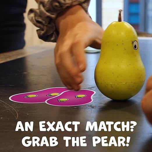 Hand reaching for a yellow pear-shaped toy with eyes next to matching purple game cards on a tabletop.
