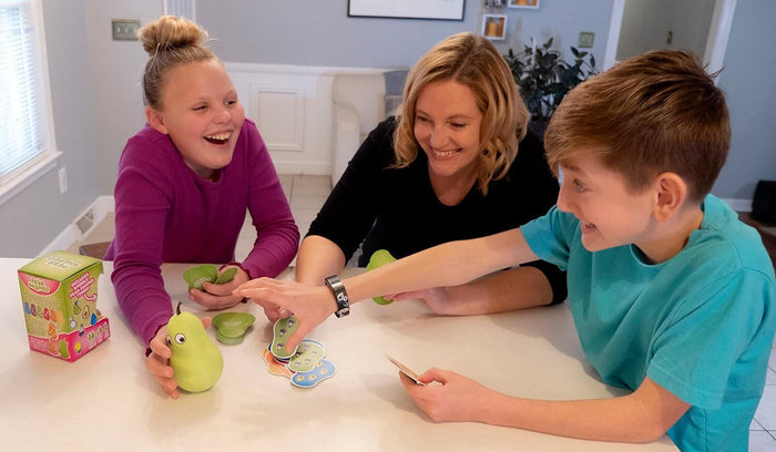 A family plays a game with pear-shaped cards, a green pear figurine, and a colorful box on a white table.