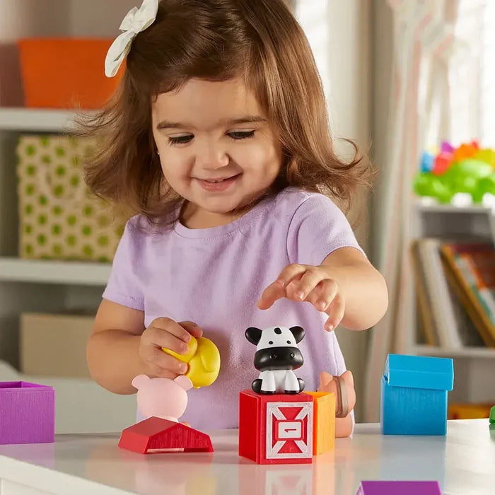 Young girl playing with colorful toy farm animal figures and stackable barn blocks on a white table.