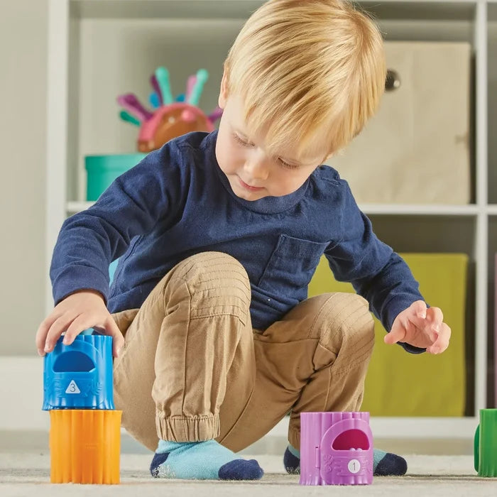 Toddler stacking colorful, numbered tree trunk toys on a rug in a playroom.