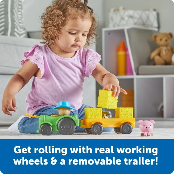 Toddler playing with a green toy tractor and yellow trailer carrying yellow hay blocks next to a small pink pig toy.