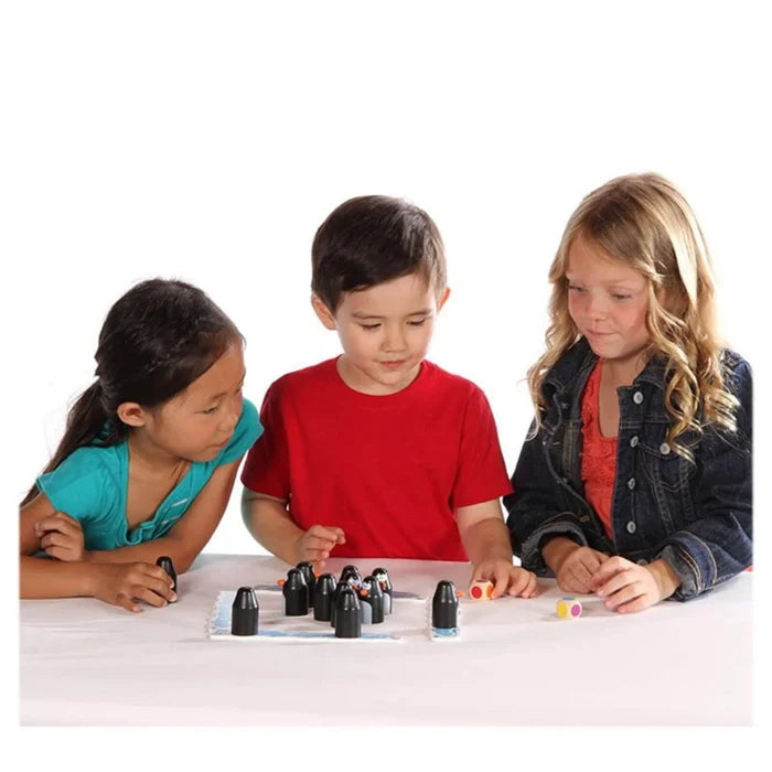 Three children playing a board game with black penguin figures and two colorful dice on a white table.