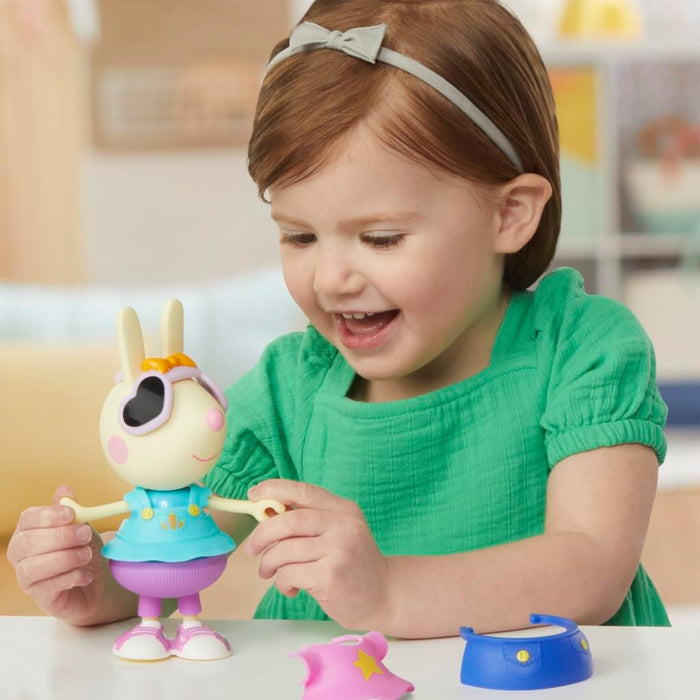 Smiling girl plays with a Rebecca Rabbit toy in a blue dress and sunglasses, with extra outfits on a table.
