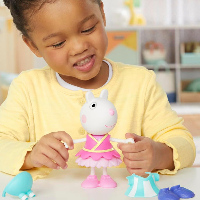A young girl plays with a Suzy Sheep figure wearing a pink tutu, with blue outfits and shoes on the table.