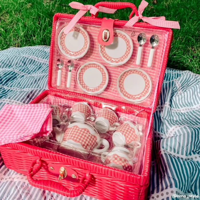 Red wicker picnic basket open to show a pink gingham toy tea set on a striped blanket in the grass.