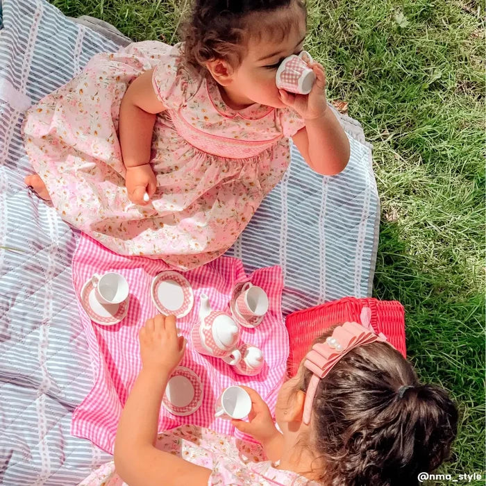 Children playing with a pink and white gingham toy tea set including a teapot, cups, and saucers on a blanket.