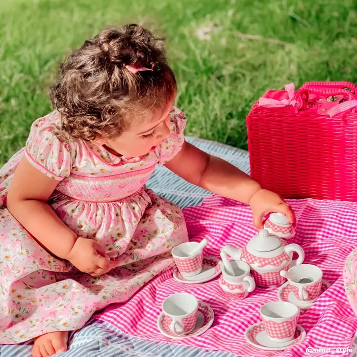 Pink gingham toy tea set with teapot and cups on a checkered blanket next to a red wicker basket and child.