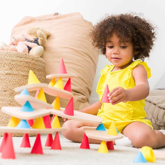 Child building a structure with red, yellow, and blue silicone cones and flat wooden boards on a white rug.