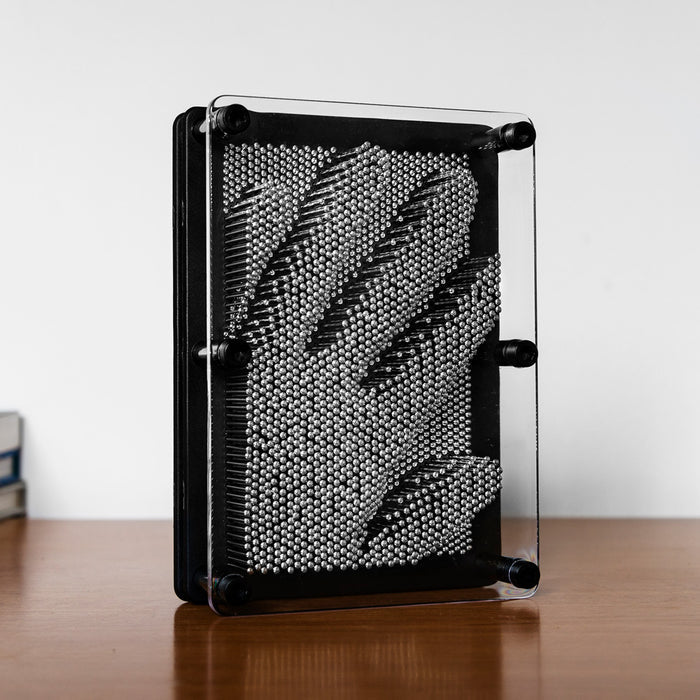 Hand impression in silver metal pins inside a black frame with clear acrylic panels, on a wooden surface.