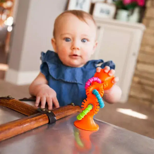 Baby playing with an orange suction toy featuring three colorful, textured loops in pink, blue, and lime green.