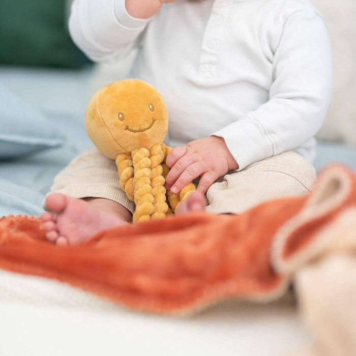 Ochre yellow plush octopus with an embroidered smiling face and coiled tentacles, shown held by a baby.