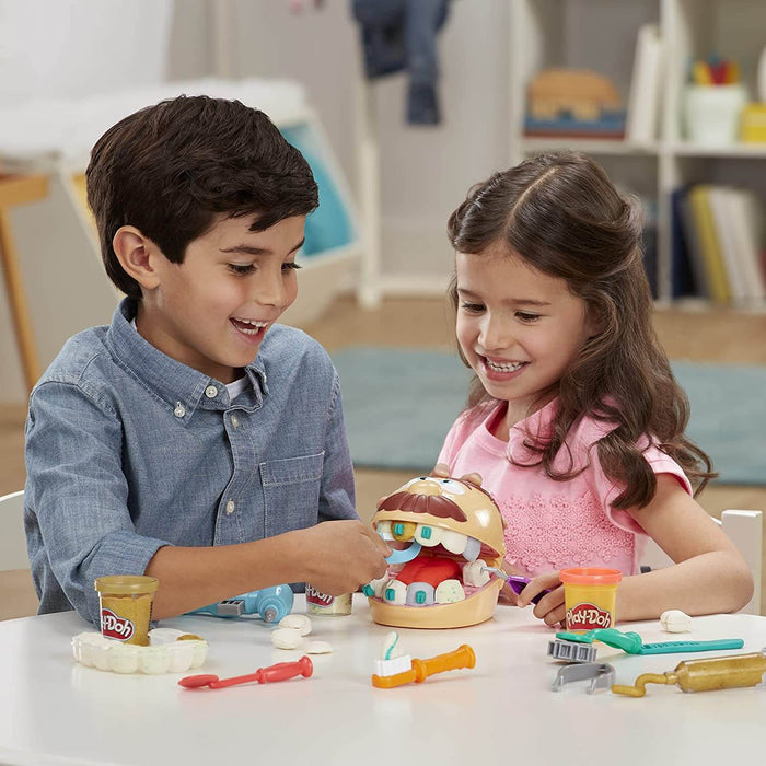 Two children use toy tools and colorful Play-Doh to fix teeth on a smiling mustache head toy at a white table.