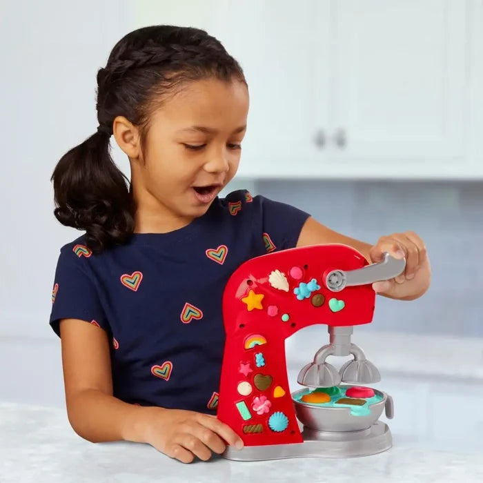 A young girl uses a red toy stand mixer decorated with colorful dough molds to create pretend treats.