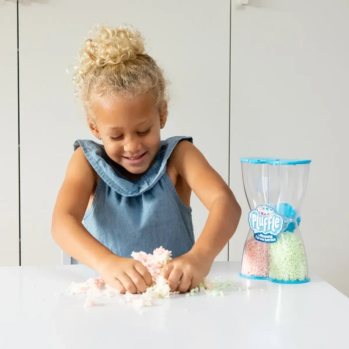 A young girl playing with pink and green foam next to a clear hourglass-shaped container on a white table.