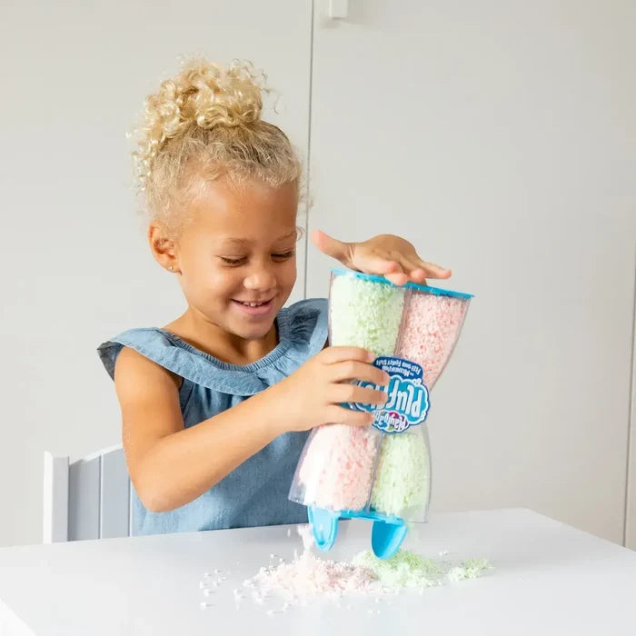 Smiling child squeezes pink and green textured foam from a clear, hourglass-shaped container onto a white table.