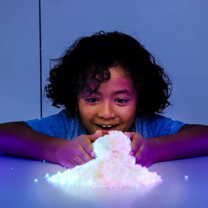 Child smiling at a glowing white mound of textured Playfoam Pluffle under purple light.