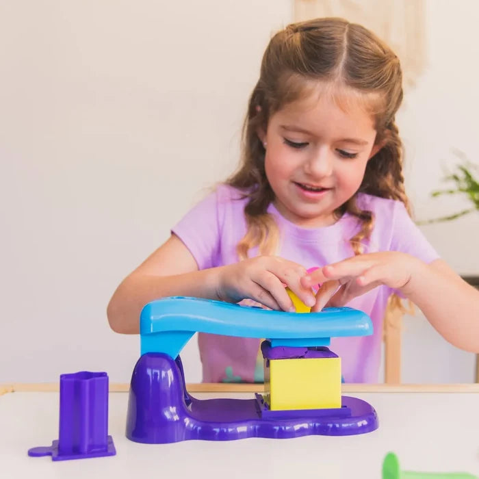 A young girl uses a blue and purple press tool to shape purple sand into a yellow square mold.