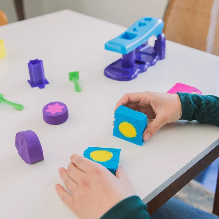 Child's hands holding two blue Playfoam Sand cubes with yellow centers, surrounded by purple and pink sand shapes.