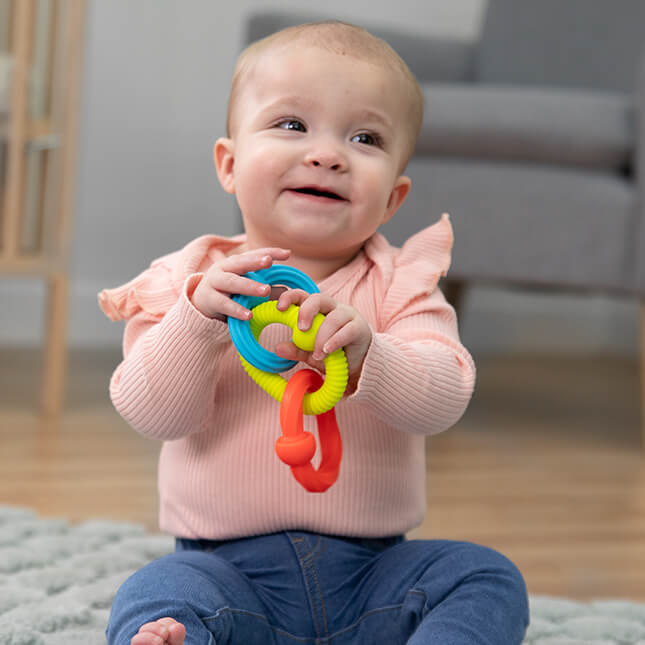 Smiling baby sitting on a rug holding three textured interlocking rings in blue, lime green, and orange.
