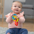 Smiling baby sitting on a rug holding three textured interlocking rings in blue, lime green, and orange.