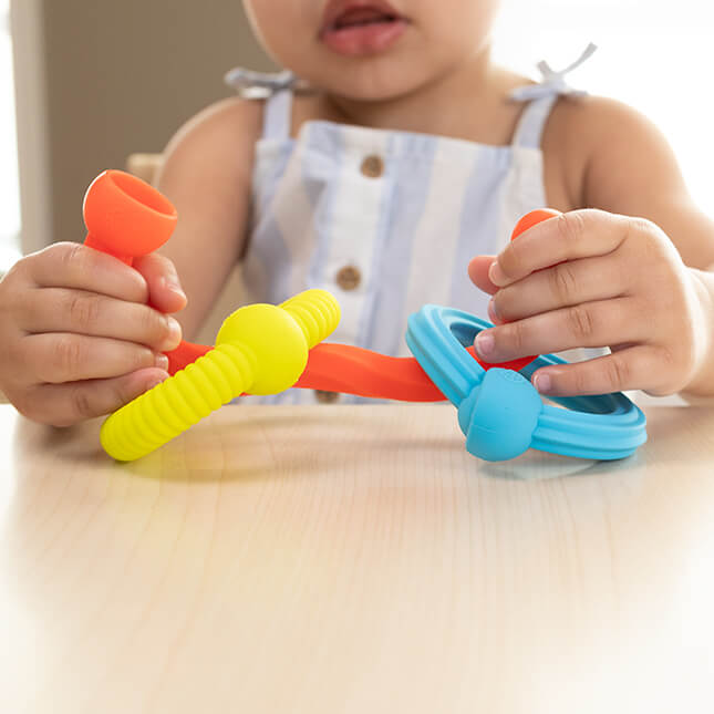 Toddler hands connecting bright, textured silicone toy pieces in orange, yellow, and blue on a table.