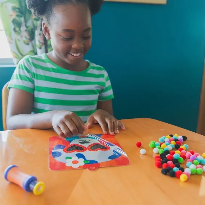 Smiling girl decorates a puppy-themed activity card with colorful mini pom poms on a wooden table.