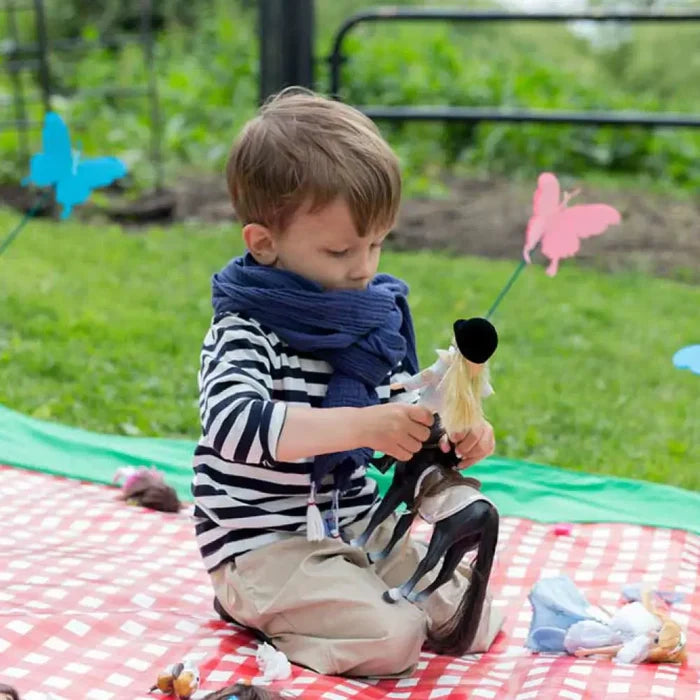 Boy plays with a blonde rider doll on a black horse toy over a red checkered picnic blanket in the grass.