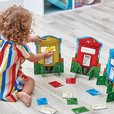 A child sorts colorful letter cards into matching red, yellow, and blue toy post boxes on a wooden floor.