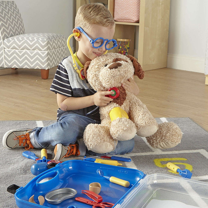 Boy wearing blue glasses uses a yellow stethoscope and blue toy medical kit to examine a stuffed dog on a rug.
