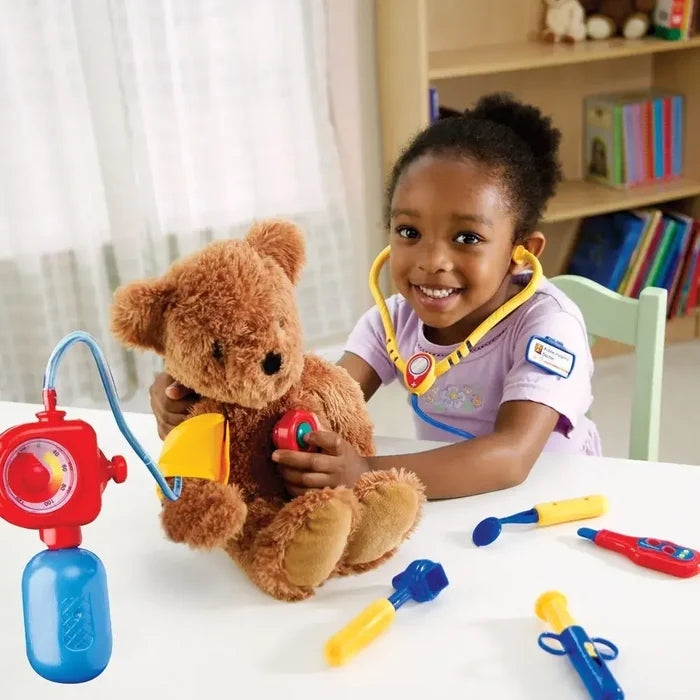 A child uses a yellow toy stethoscope and red blood pressure monitor to examine a brown teddy bear on a white table.
