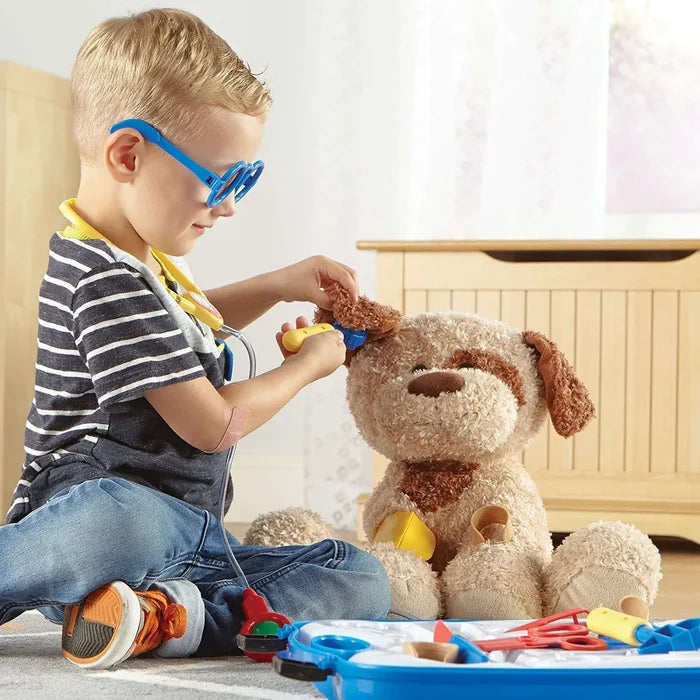 Boy in blue toy glasses uses a yellow tool on a stuffed dog's ear. An open blue kit with colorful tools sits nearby.