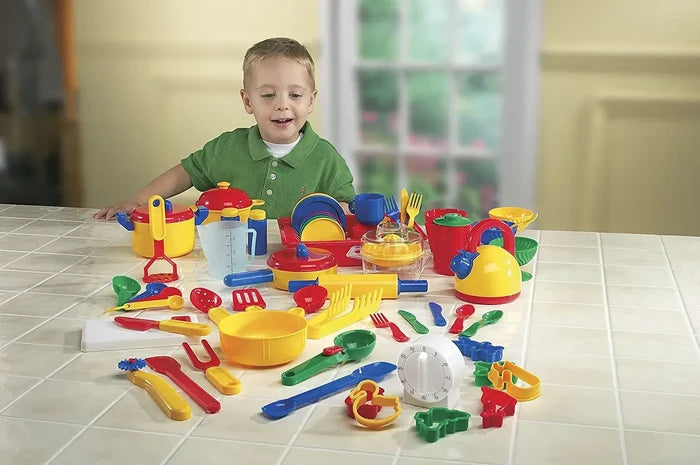 Smiling boy behind an array of vibrant red, yellow, and blue plastic toy kitchen accessories on a tiled counter.
