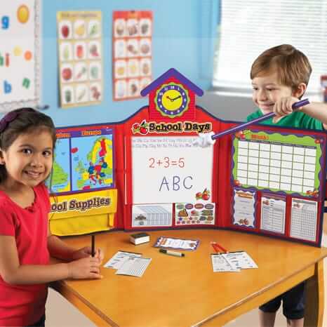 Two children play with a red schoolhouse playset featuring a whiteboard, clock, map, and toy classroom accessories.