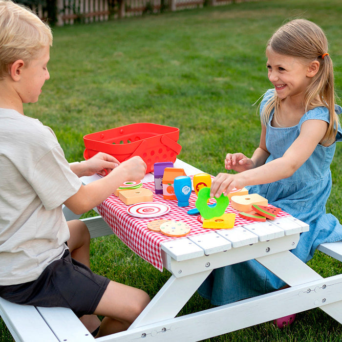 Two children play with a red toy picnic basket and colorful wooden food on a white table with a checkered cloth.