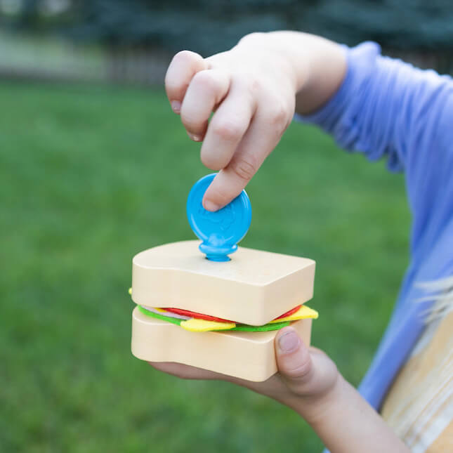 Child's hands holding a wooden toy sandwich with colorful felt layers and a blue circular pick.
