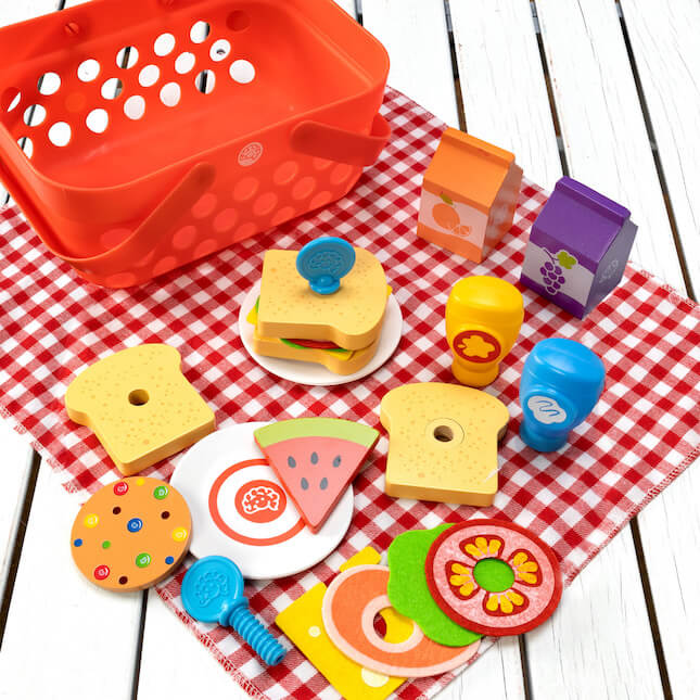 Red toy picnic basket with wooden play food: sandwich, fruit, and juice boxes on a red and white checkered cloth.