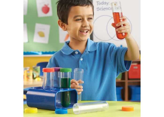 Smiling boy holds a jumbo test tube with red liquid, with more colorful tubes in a blue plastic rack on a table.