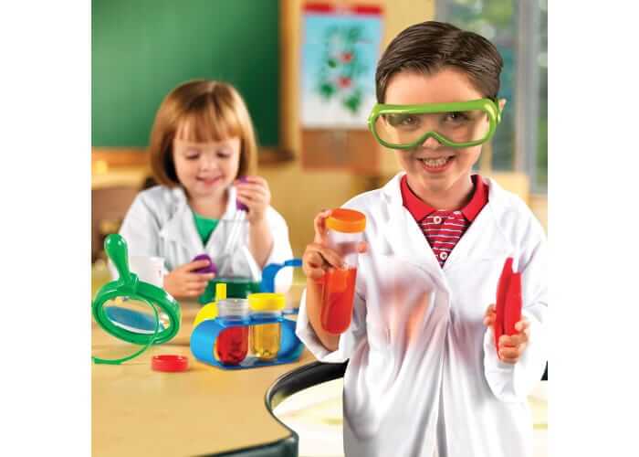 Two children in lab coats and goggles playing with colorful test tubes, a magnifying glass, and tweezers.