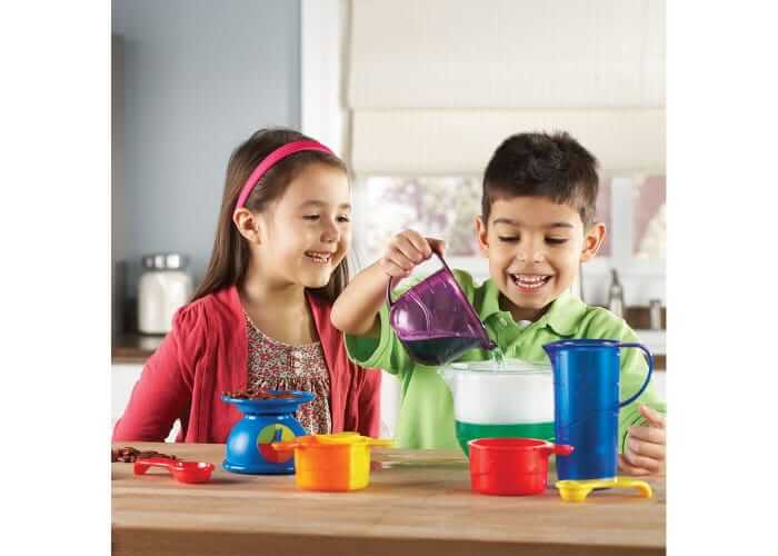 Two children happily use colorful red, blue, and yellow measuring cups, spoons, and a toy scale to pour liquids.