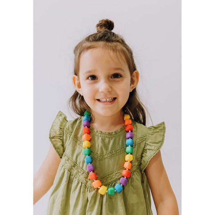 A young girl wearing a chunky chewable necklace with large, rounded square beads in vibrant rainbow colors.
