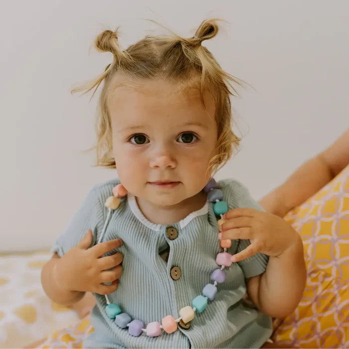 Toddler wearing a chewable necklace featuring pastel-colored hexagonal beads in pink, purple, blue, and green.