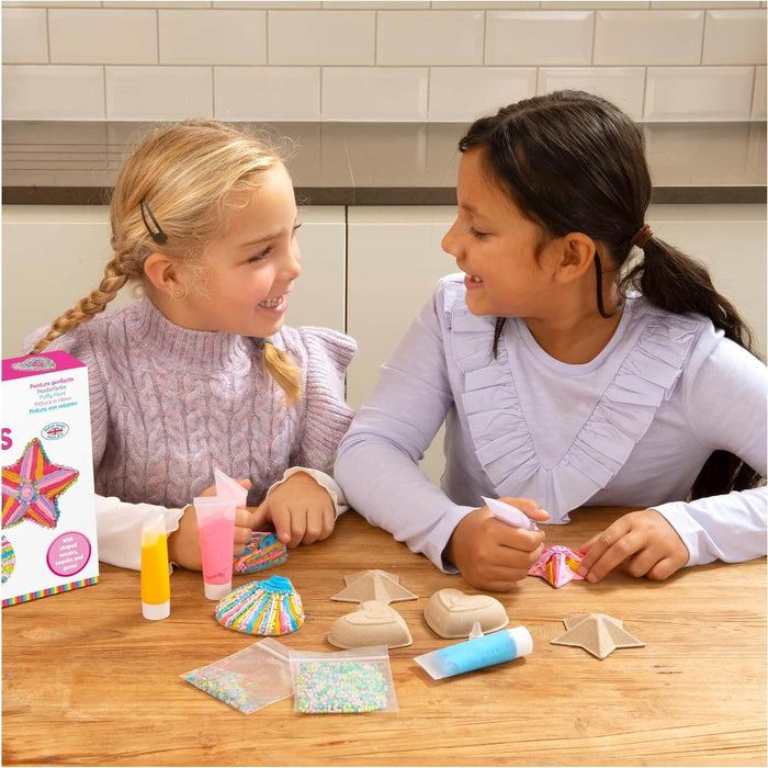 Two girls decorate cardboard shapes using colorful puffy paint tubes and gems at a wooden table.