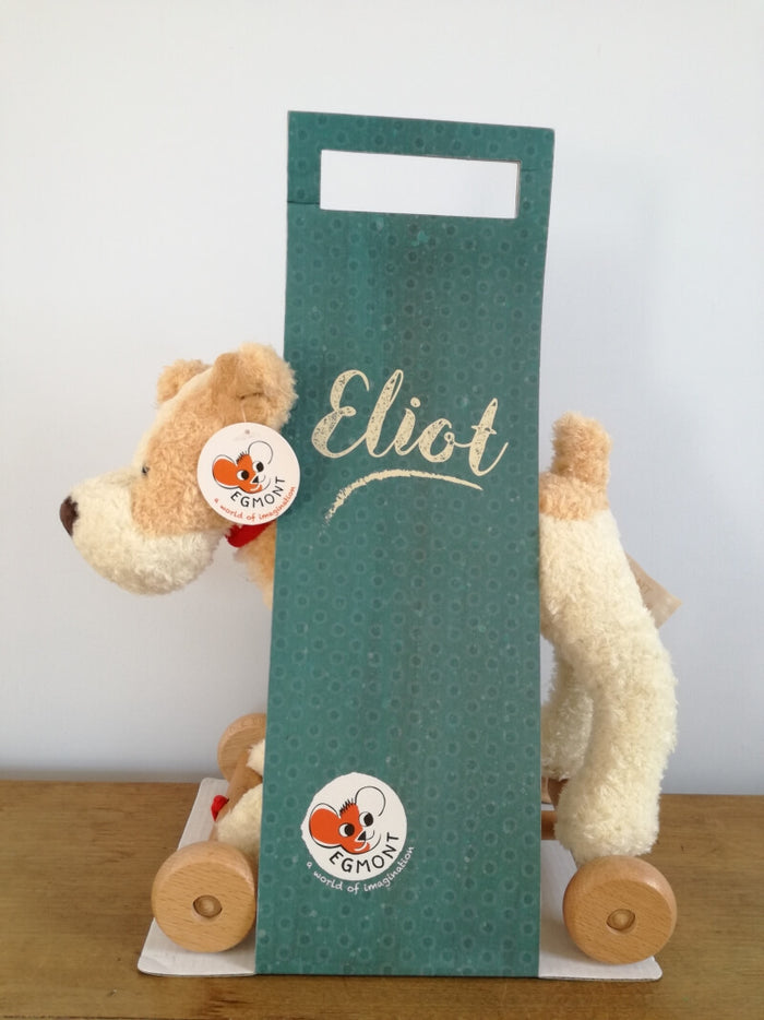 Plush cream and tan dog on wooden wheels, inside a tall teal cardboard sleeve printed with Eliot in white script.