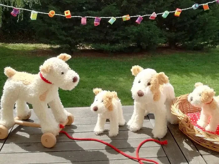 White and tan plush pull-along dog on wooden wheels with red cord, on a deck with other matching stuffed toy dogs.
