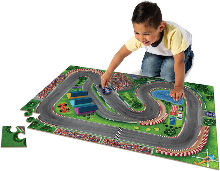 A boy plays with a large race track floor jigsaw puzzle and wooden toy cars featuring bright green and grey colors.