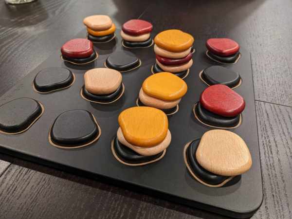 Smooth pebble-shaped game pieces in red, yellow, black, and tan stacked on a dark grey 4x4 grid game board.