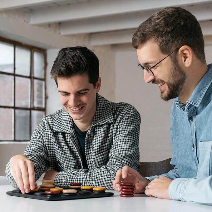 Two men playing a strategy game with smooth tan, yellow, and red wooden stones on a black grid board.