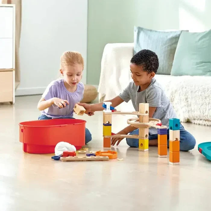 Two children building a wooden marble run with colorful blocks and tracks next to a large red storage bucket.