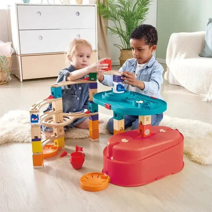 Two children building a colorful wooden marble run with tracks, blocks, and a red storage bucket base.